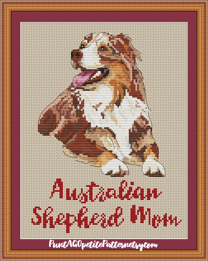 May include: Cross-stitch pattern of an Australian Shepherd dog sitting with its tongue out. The pattern is framed in a brown and red frame. The text "Australian Shepherd Mom" is below the dog and the website "PuntAGOpetitePatternetsy.com" is below the text.