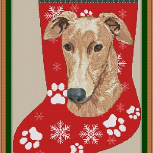 Puede incluir: Una media de Navidad roja con la cara de un perro Greyhound blanco y la palabra "GREYHOUND" en la parte superior. La media está decorada con copos de nieve blancos y huellas de patas.