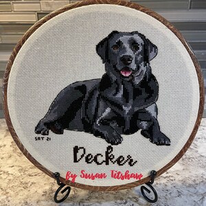 May include: A black Labrador Retriever cross-stitch in a brown wooden embroidery hoop with the name "Decker" and the artist's name "by Susan Titshaw" in black lettering.