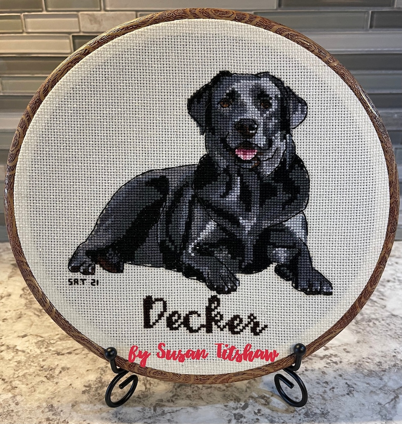 May include: A black Labrador Retriever cross-stitch in a brown wooden embroidery hoop with the name "Decker" and the artist's name "by Susan Titshaw" in black lettering.