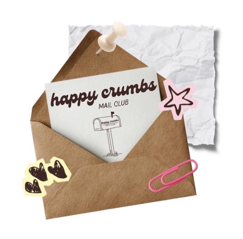 Pu&ograve; includere: Una busta marrone con una carta bianca che recita "happy crumbs MAIL CLUB" e un disegno di una cassetta postale. La busta &egrave; fissata con una puntina e decorata con adesivi, tra cui una stella rosa, cuori e una graffetta rosa.