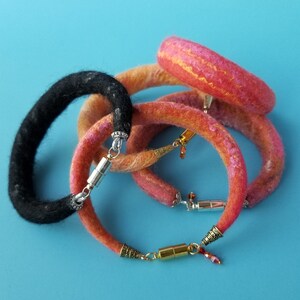 May include: Four felted wool bracelets in black, orange, and pink with silver and gold accents. The bracelets are stacked on top of each other.