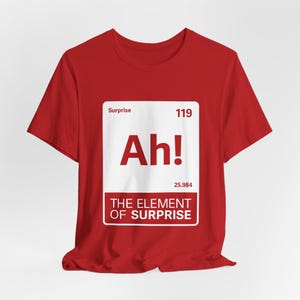 Peut inclure: Un t-shirt rouge à manches courtes avec un graphique rectangulaire blanc ressemblant à un élément du tableau périodique. Le graphique affiche le texte Surprise, 119, Ah!, 25.984 et THE ELEMENT OF SURPRISE en lettres rouges et blanches.