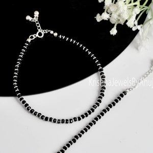 Op de afbeelding: Twee zilveren enkelbandjes met zwarte kralen. De enkelbandjes hebben een zilveren ketting met een kleine bal aan het einde.
