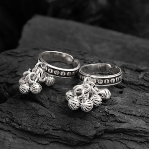 May include: Two silver toe rings with dangling bell charms. Each ring has a patterned band and a cluster of small, round bells. The rings are displayed on a dark, textured surface.