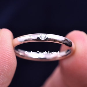 Puede incluir: Un anillo plateado pulido con una pequeña gema transparente engastada en la banda. El anillo se sostiene entre dos dedos sobre un fondo oscuro. El texto "Krishna JewelsByAnuj" es visible.