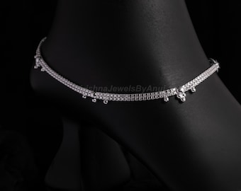 May include: A silver anklet with a delicate design, featuring small dangling accents. The anklet is worn on a dark surface, showcasing its intricate details. The text "KrishnaJewelsByAnu" is visible.