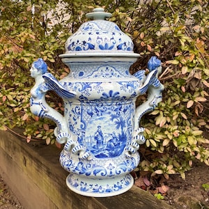 May include: A large, ornate blue and white ceramic vase with a lid. The vase features detailed floral and landscape designs, with handles adorned with sculpted figures. The overall height is approximately 24 inches.