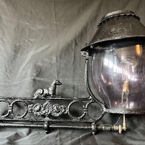 May include: An antique black metal outdoor lamp with a decorative dog figure and floral accents. The lamp features a large, clear glass globe and a dark, conical shade. The lamp is designed to be mounted on a wall.