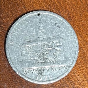 May include: A silver-colored round medal with a building illustration and the text "W. MITCHELTREE 1910". The medal has a small hole at the top, suggesting it could be hung. The building has a detailed architectural design.