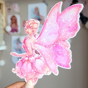 May include: A pink fairy sticker with glitter details. The fairy is wearing a light pink dress and has large, detailed wings. The sticker is held by a hand with manicured nails. The fairy is a whimsical design.