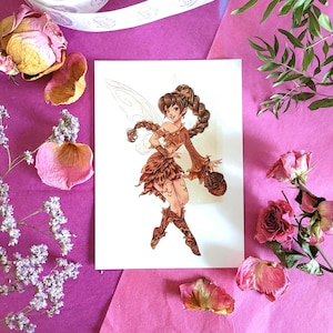 Peut inclure: Illustration d&eacute;taill&eacute;e d'une f&eacute;e aux cheveux bruns et aux ailes, v&ecirc;tue d'une tenue marron, tenant un petit sac. L'&oelig;uvre d'art est sur une carte blanche, entour&eacute;e de roses s&eacute;ch&eacute;es, de p&eacute;tales et de verdure sur fond rose.
