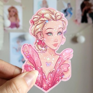 May include: A pink sticker depicting a woman with blonde hair and blue eyes. The sticker has a holographic effect with star-shaped sparkles. A butterfly is on the chest of the woman. The sticker is held by a hand.
