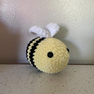 May include: A handmade, plush bee toy. The bee features a yellow body with black stripes, black eyes, and white wings. The toy is made of soft, textured yarn, giving it a cuddly appearance. This item is a cute, decorative piece.