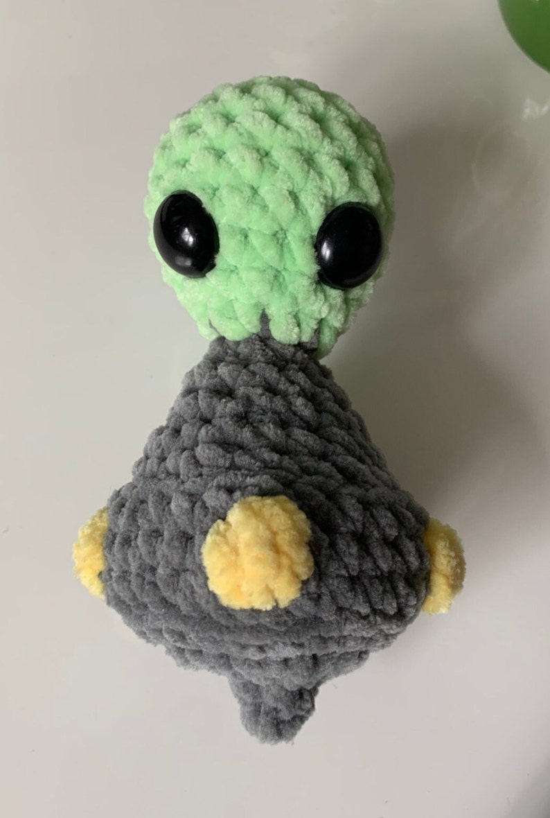 Pu&ograve; includere: Un giocattolo di peluche all'uncinetto verde e grigio con occhi neri e dettagli gialli. Il giocattolo ha la forma di un piccolo alieno con una testa rotonda e un corpo triangolare.