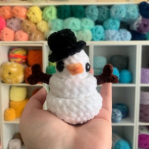May include: A handmade white snowman with a black top hat, brown arms, and an orange nose. The snowman is made of soft, textured yarn. The background features shelves filled with colorful yarn.