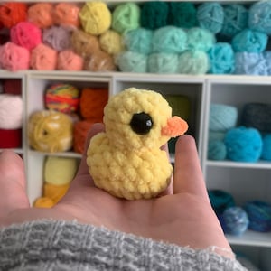 May include: A handmade yellow crochet duck with a black eye and orange beak. The small plush duck is held in a hand, with a background of colorful yarn skeins on shelves. The duck is a cute, whimsical craft item.