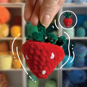 Puede incluir: Una fresa roja de crochet con hojas verdes peludas. La fresa está siendo sostenida por una mano y está enfocada. El fondo está desenfocado y muestra un estante con ovillos de lana de colores.