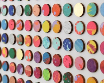 May include: A wall art piece featuring numerous wooden circles, each with a unique color and paint splatter design. The circles are arranged in a grid pattern, showcasing a variety of colors including red, blue, yellow, and gold. The artwork has a modern, abstract aesthetic.