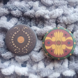 Könnte beinhalten: Zwei runde dekorative Magnete. Einer ist braun mit einem goldenen Schlangenmuster und einem zentralen Kreis. Der andere ist grün, rot und goldfarben mit einem floralen Muster. Beide liegen auf einer grau strukturierten Oberfläche.