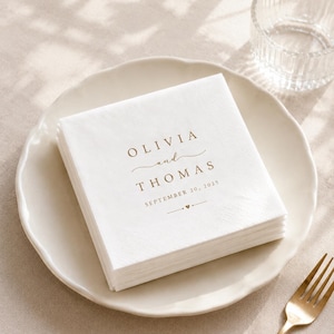 May include: A stack of square white paper napkins featuring gold-coloured text that reads OLIVIA and THOMAS, SEPTEMBER 20, 2025, with a small heart icon. The napkins are placed on a white plate next to a gold-coloured fork and a clear glass.
