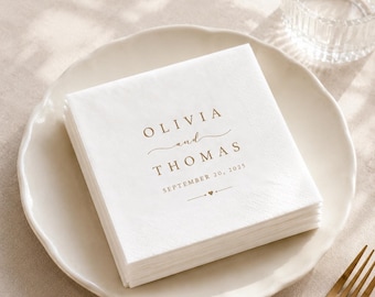 May include: A stack of square white paper napkins featuring gold-colored text that reads OLIVIA and THOMAS, SEPTEMBER 20, 2025, with a small heart icon. The napkins are placed on a white plate next to a gold-colored fork and a clear glass.