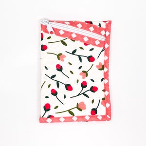 May include: A rectangular, quilted pouch with a white zip. The pouch has a white background with a floral pattern of red buds, pink flowers, and green leaves. The edges are trimmed with a pink and white diamond pattern.