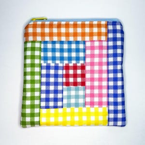 May include: A rectangular zippered pouch featuring a patchwork design of multi-colored gingham fabric squares. The pattern includes orange, green, blue, red, light blue, pink, and yellow checkered sections with a light blue zipper and yellow pull.