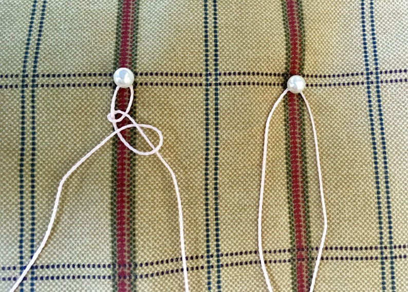May include: Two white pearls are strung on white thread. The thread is tied in a knot on the left pearl and is straight on the right pearl.