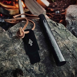 Puede incluir: Un iniciador de fuego negro con una correa de cuero y un percutor de metal. El iniciador de fuego está sobre una roca gris.