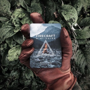 Peut inclure: Une main gantée de cuir marron tient une carte avec le texte "Firecraft Mini-Guide" et un graphique d'un feu de camp. La carte porte également le texte "Bushlore Handcrafted Survival".