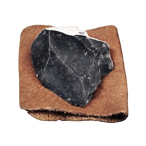 May include: A dark grey, irregularly shaped flint stone with a smooth, glossy surface and visible white mineral inclusions, resting on a square piece of textured, light brown leather with a visible grain and a folded bottom edge.