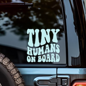 May include: A black SUV with a "Tiny Humans On Board" decal in light blue lettering on the rear window. The decal is in a retro font. The vehicle has a spare tire and a red taillight.