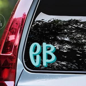 May include: A teal-colored vinyl decal with the letters "GB" on a car window. The letters have a 3D effect with a shadow. The decal is on the rear window of a vehicle, with a blurred background of trees.