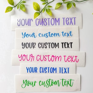 May include: Six rectangular adhesive vinyl decals featuring the phrase "YOUR CUSTOM TEXT" in various fonts and colors, including light purple, blue, black, pink, dark blue, and green, arranged in a vertical stack on a white surface.