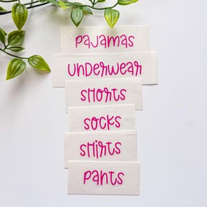 May include: A set of white rectangular labels with pink text featuring the words pajamas, underwear, shorts, socks, shirts, and pants. The labels are arranged in a vertical stack, and a green leafy vine is positioned at the top left corner.