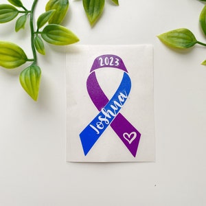 May include: A blue and purple ribbon with the name "Joshua" and the year "2023" written on it. A small purple heart is in the bottom right corner of the ribbon.