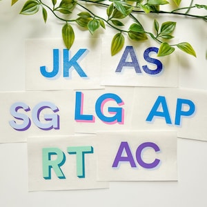 May include: A set of eight colorful, 3D letter stickers. The letters are in various colors, including blue, pink, purple, and green. The letters spell out "JK AS SG LG AP RT AC".