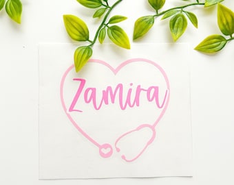 May include: A light pink vinyl decal featuring the name Zamira written in a cursive font inside a heart shape formed by a stethoscope outline. The stethoscope includes a small heart icon at the bottom, presented on a white square backing.