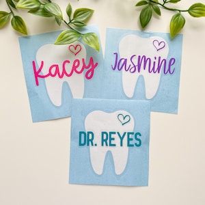 May include: Three square light blue stickers featuring a white tooth graphic and a small heart. The stickers display the names Kacey in pink, Jasmine in purple, and the text DR. REYES in teal, each written in a distinct font style.