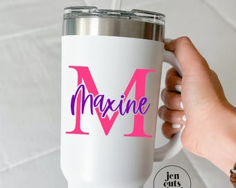 May include: A white stainless steel tumbler with a handle, a clear lid, and a straw. The tumbler features a large hot pink letter M with the name Maxine written in purple script across it. A small circular logo reads jen cuts vinyl.
