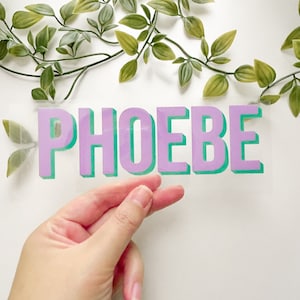 Könnte beinhalten: Ein transparenter Vinyl-Aufkleber mit dem Namen "PHOEBE" in großen, 3D-artigen Buchstaben. Die Buchstaben sind lila mit grünem Umriss.