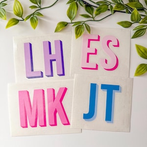 May include: Four square stickers with initials in a 3D effect. The initials are LH in purple, ES in pink, MK in pink, and JT in blue. Green leafy vines are in the background.