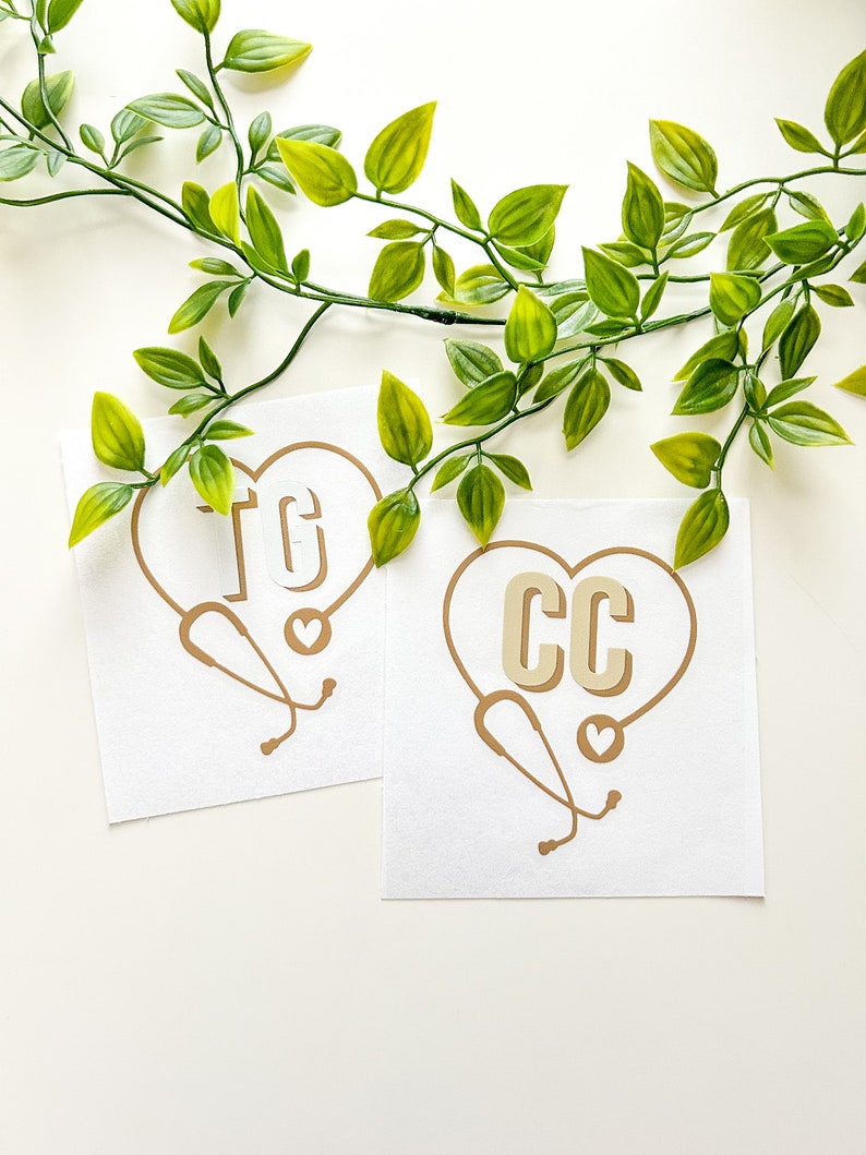 Peut inclure: Deux cartes blanches avec des motifs en forme de c&oelig;ur dor&eacute;s repr&eacute;sentant les initiales &laquo;&nbsp;TG&nbsp;&raquo; et &laquo;&nbsp;CC&nbsp;&raquo; avec un st&eacute;thoscope et un petit c&oelig;ur. Les cartes sont sur une surface blanche avec une vigne feuillue verte.