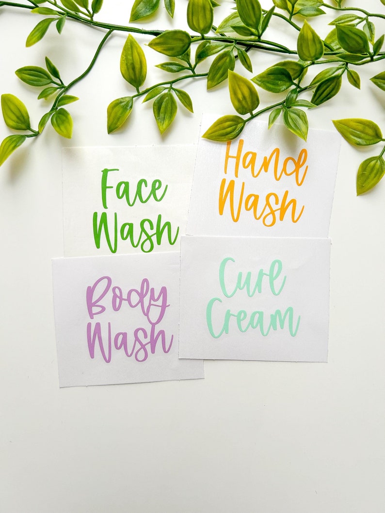 May include: Four white rectangular adhesive labels featuring colorful script text. The labels display the phrases Face Wash in green, Hand Wash in orange, Body Wash in purple, and Curl Cream in light green, arranged in a grid-like layout.