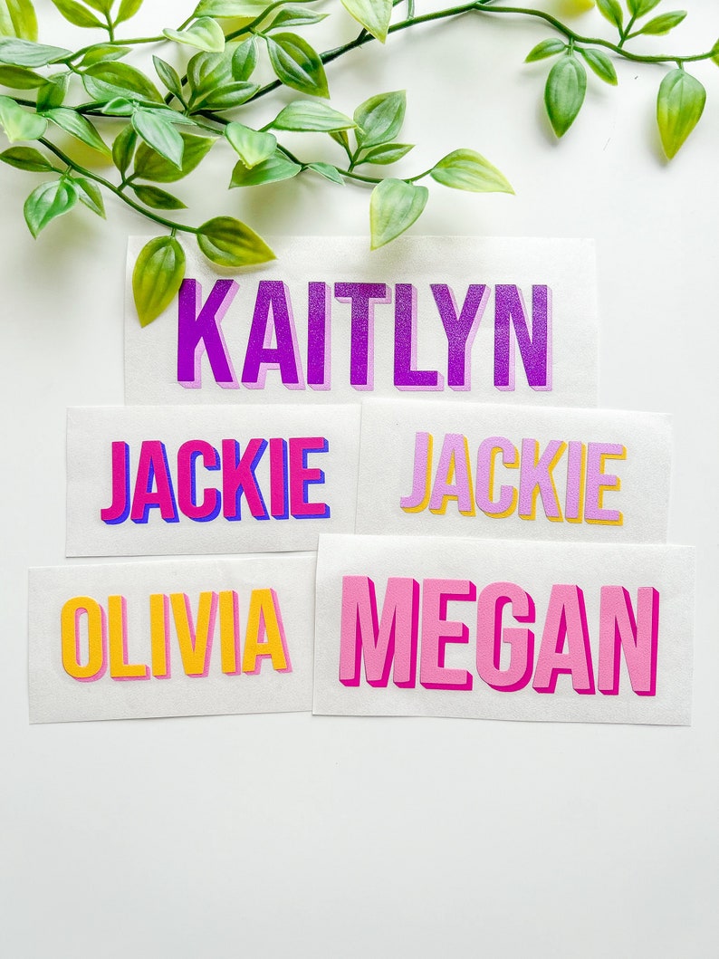 Pu&ograve; includere: Cinque adesivi rettangolari in vinile bianco con i nomi KAITLYN in viola, JACKIE in rosa e blu, JACKIE in viola chiaro e giallo, OLIVIA in giallo e rosa, e MEGAN in rosa, tutti visualizzati con un carattere in grassetto in stile 3D.