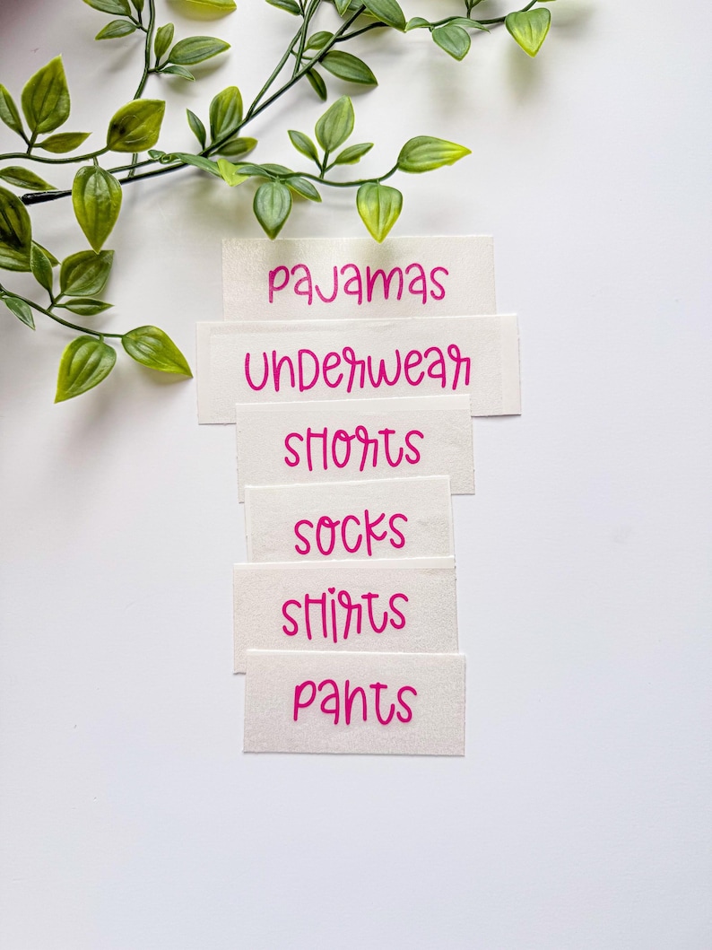 May include: A set of white rectangular labels with pink text featuring the words pajamas, underwear, shorts, socks, shirts, and pants. The labels are arranged in a vertical stack, and a green leafy vine is positioned at the top left corner.