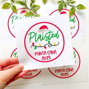Puede incluir: Una pegatina cuadrada blanca con un borde circular rojo, el texto verde Plaisted, un gorro de Pap&aacute; Noel rojo, una cadena de luces navide&ntilde;as de colores y el texto rojo PUNTA CANA 2023. El dise&ntilde;o est&aacute; impreso en varias hojas de papel cuadradas.