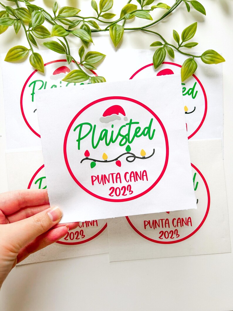 Puede incluir: Una pegatina cuadrada blanca con un borde circular rojo, el texto verde Plaisted, un gorro de Pap&aacute; Noel rojo, una cadena de luces navide&ntilde;as de colores y el texto rojo PUNTA CANA 2023. El dise&ntilde;o est&aacute; impreso en varias hojas de papel cuadradas.