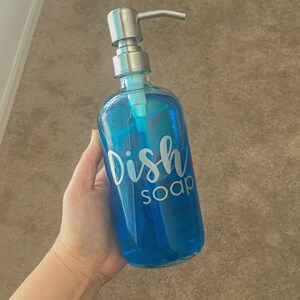 May include: A clear blue glass soap dispenser with a silver-toned metal pump mechanism. The front of the bottle features the white text Dish soap in a cursive and sans-serif font combination. The bottle is filled with blue liquid soap.